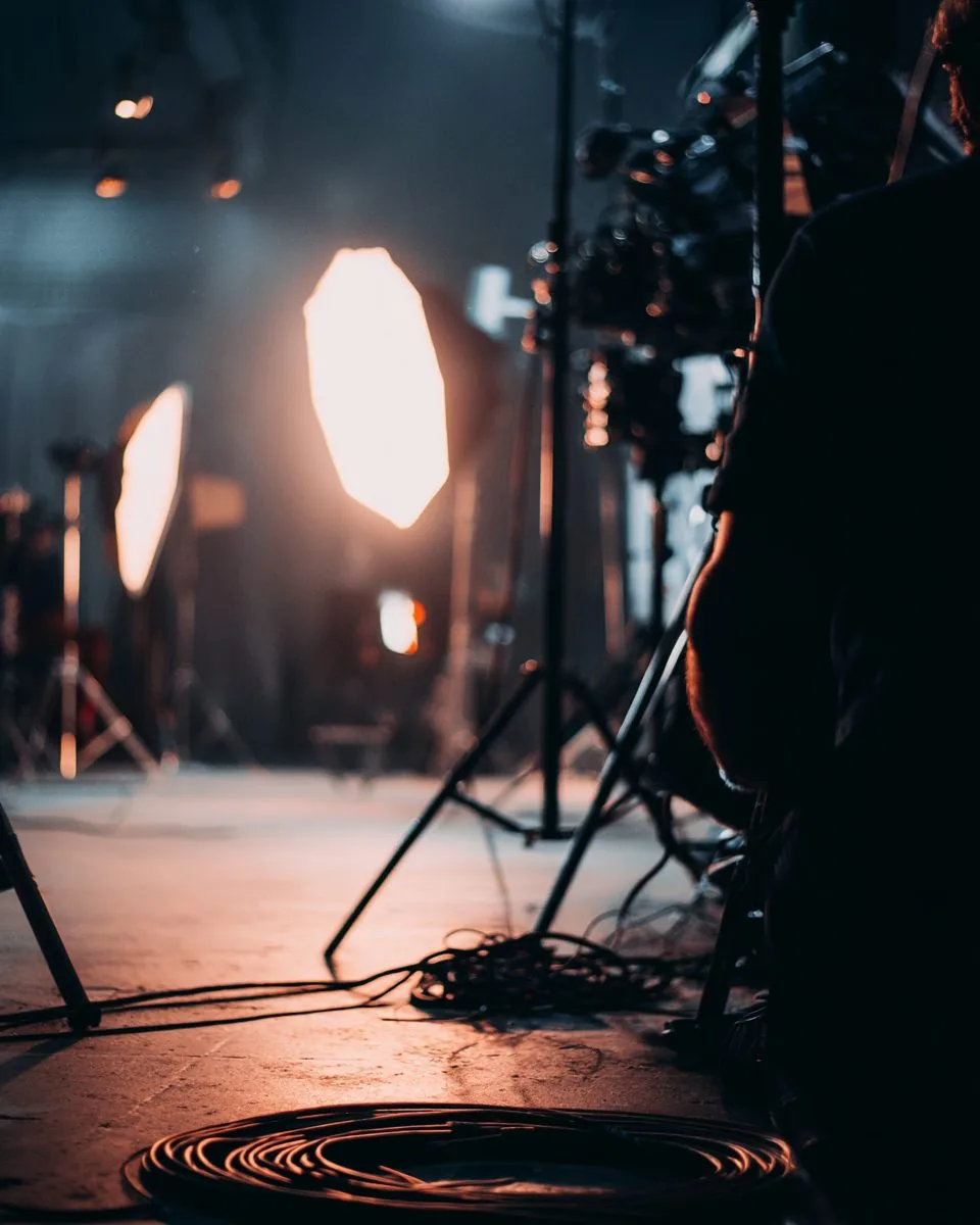 A film set between takes: softbox lighting out of focus, coiled cables and a crew member in silhouette.